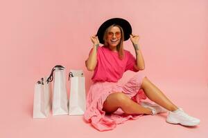 Pretty smiling woman in  spring trendy outfit posing with white shopping bags in studio over pink background. photo