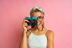 Laughing blond woman  holding retro camera and posing on pink background. Travel and summer vacation concept. photo