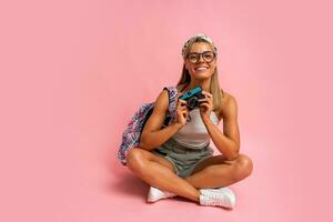 Beautiful tourist woman with camera and back pack sitting on floor in studio on pink background. Vacation mood. photo