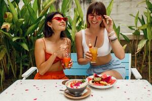 Two pretty friends, stylish girls spending  leisure time tropical cafe, enjoying tasty food, smoothie bowls, lemonade and fruit plate. Party and vacation mood. Trendy sunglasses. photo