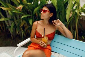 Stunning tan  woman  in retro sunglasses relaxing in tropical beach club in stylish red top and orange shorts. Drinking tasty lemonade.  Vacation and holidays. photo