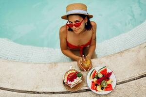 Confident  brunette tan woman in red cat eyes sunglasses   relaxing in   pool  with plate of exotic fruits  during tropical vacation.  Stylish tattoo. photo