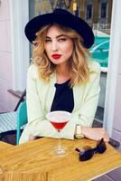 Stylish woman in casual summer outfit in black hat sitting in modern cafe and drinking Cocktail. photo