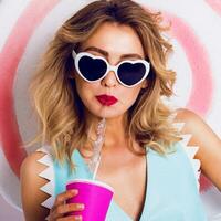 Pretty girl in sunglasses with beautiful skin and lips, posing in studio, drinking fruit juice or cocktail. Wearing vintage heart sunglasses, stylish blue leather photo