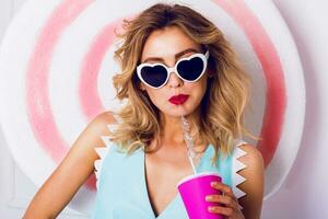 Pretty girl in sunglasses with beautiful skin and lips, posing in studio, drinking fruit juice or cocktail. Wearing vintage heart sunglasses, stylish blue leather photo