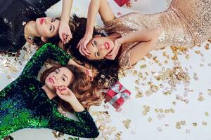 Top view on three gorgeous girls lying on the floor, celebrating new year or birthday party. Wearing luxury sequins dress  and jewelry. Golden shining confetti , red gift boxes. photo