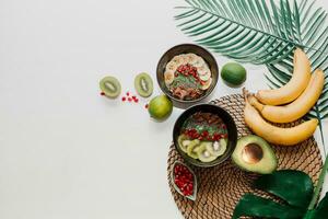 Healthy food concept. Top view on table with smoothie bowls.  Plate topped with kiwi, granola, garnet, chia, avocado. Tropical leaves on background. Copy space. photo