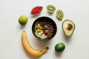 Essentials for cooking smoothie bowl. Blake plate topped with Kiwi, banana, pomegranate seeds, lime, granola, chia seeds. Healthy breakfast.  Round composition on white background. photo