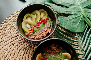 Healthy food concept. Top view on table with smoothie bowls.  Plate topped with kiwi, granola, garnet, chia, avocado. Tropical leaves on background. Copy space. photo