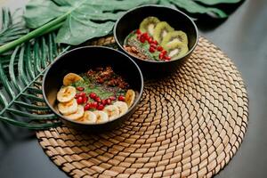 Healthy food concept. Top view on table with smoothie bowls.  Plate topped with kiwi, granola, garnet, chia, avocado. Tropical leaves on background. Copy space. photo