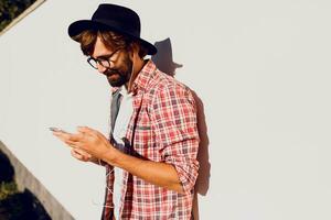 Half length portrait of bearded male with retro style using cell telephone while standing in urban white wall ,  chatting on smart phone during walking in spring day. Copy space for text. photo