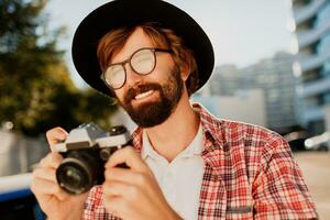 cerca arriba retrato de sonriente hipster barba hombre utilizando retro película cámara, haciendo fotos mientras de viaje. urbano antecedentes. elegante primavera atuendo. vistiendo negro sombrero, a cuadros camisa .