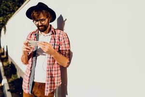 Half length portrait of bearded male with retro style using cell telephone while standing in urban white wall ,  chatting on smart phone during walking in spring day. Copy space for text. photo