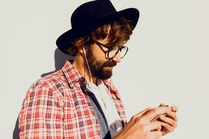 portrait of bearded male with retro style using cell telephone while standing in urban white wall ,  chatting on smart phone during walking in spring day. Copy space for text. photo