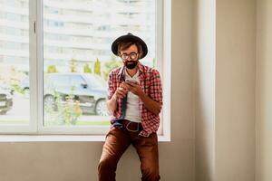 barbado hombre en brillante a cuadros camisa instalando nuevo móvil solicitud en teléfono inteligente dispositivo y escuchando música. hipster estilo. foto