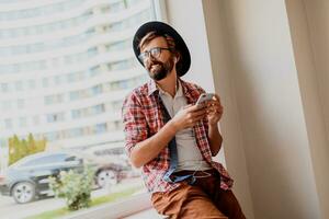 Handsome  hipster male with beard   in sunglasses texting messages via smartphone and blogging in social networks sharing multimedia  near copy space for advertising. Sitting o near window. photo