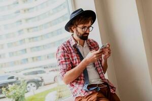 Handsome  hipster male with beard   in sunglasses texting messages via smartphone and blogging in social networks sharing multimedia  near copy space for advertising. Sitting o near window. photo