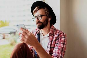 elegante barbado hombre en brillante a cuadros camisa instalando nuevo móvil solicitud en teléfono inteligente dispositivo y escuchando música. hipster estilo. foto