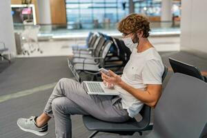 New normal and social distance concept. Man tourist wearing mask using laptop computer searching airline flight status and sitting with distance during corona virus. photo