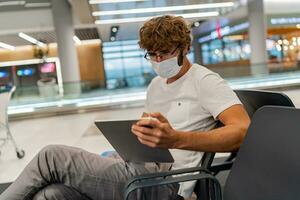 Man in respirator mask is waiting next plane at the airport and using  tablet. Coronavirus COVID-19 concept. photo