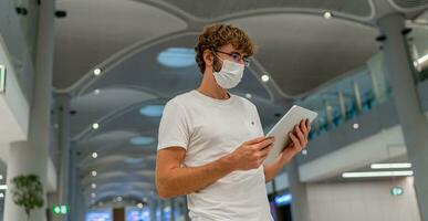 Man in respirator mask is waiting next plane at the airport and using  tablet. Coronavirus COVID-19 concept. photo