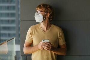 blanco barbado adulto hombre utilizando teléfono inteligente mientras vistiendo quirúrgico máscara en un industrial pared. salud, epidemias, social medios de comunicación. foto