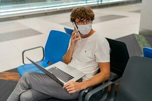 New normal and social distance concept. Man tourist wearing mask using laptop computer searching airline flight status and sitting with distance during corona virus. photo