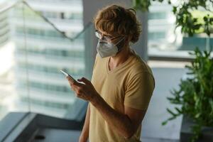 White bearded adult man using smartphone while wearing surgical mask on an industrial wall. Health, epidemics, social media. photo