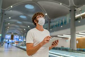 Man in respirator mask is waiting next plane at the airport and using  tablet. Coronavirus COVID-19 concept. photo