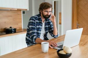European man  works from home. He sits in the kitchen at the table and uses a laptop computer and speaks on the phone. photo