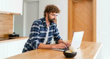 europeo hombre con barba en a cuadros camisa trabajando en ordenador portátil en el cocina a hogar. foto