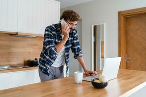 hombre con barba hablando por teléfono y utilizando ordenador portátil en el cocina. trabajando a hogar. foto