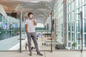 Man wearing face mask  in airport and  talking by smartphone . Health and travel concepts during coronavirus pandemic. Full lenght. photo