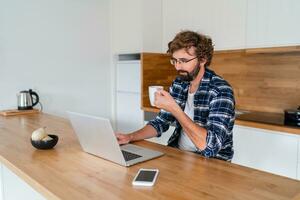 European  man with beard   in checkered shirt  smiling and working on laptop in the kitchen at home. photo