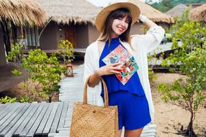 Sunny summer portrait of cute brunette woman in straw hat and trendy  boho bag walking in tropical villa in Thailand. photo