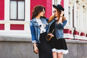 Close up  summer lifestyle portrait of two best friends walking  and talking  outdoor on the street in city center. Wearing stylish jeans jacket , dress, sunglasses. photo