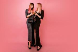 Full length studio image of celebrating women in party dress drinking shampagne and have great time together. Pink background. Happy emotions. photo