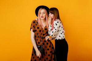 Two gossip women standing over yellow background in stylish vintage outfit. Fashionable blond and brunette females talking in studio. photo