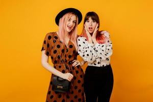 Fashionable studio shot of two stylish women with pink hairs posing over yellow background.  Ecstatic emotions. Shopping and friendship concept. photo