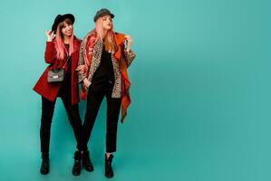 Full height image of two happy cheeky girls , best friends posing  on blue background. Wearing stylish casual   fur coat with leopard print  and hat. Space for text. photo