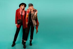 Full height image of two happy cheeky girls , best friends posing  on blue background. Wearing stylish casual   fur coat with leopard print  and hat. Space for text. photo