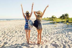 Outdoor shot of cheerful young female friends  walking together on a beach at sunset. Two attractive women enjoying a holiday on the sea shore. Wearing trendy  summer outfit, backpacks. Back. photo