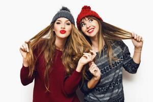 Two  cheerful young friends in perfect mood having time together on white isolate background. Wearing   trendy winter sweater and knitted red and grey hat . Positive and friendship concept. photo