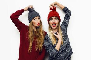 Two  cheerful young friends in perfect mood having time together on white isolate background. Wearing   trendy winter sweater and knitted red and grey hat . Positive and friendship concept. photo