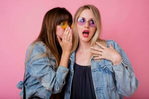 Two emotional girls gossip up on pink background . Blonde woman with surprise face listening her brunette friend. photo