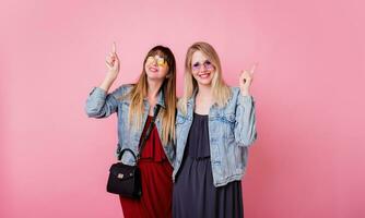Lifestyle studio image of two cheerful cute women,  best friends having great time together , posing over pink background . Wearing jeans jackets and elegant dress . photo