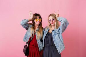 Lifestyle studio image of two cheerful cute women,  best friends having great time together , posing over pink background . Wearing jeans jackets and elegant dress . photo