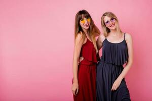 Couple of smiling women posing on pink background. Two smiling friends  hugging and looking on camera. Stylish similar dress. photo