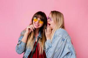 Two emotional girls gossip up on pink background . Blonde woman with surprise face listening her brunette friend. photo
