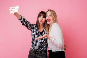 dos bonito muchachas haciendo yo retrato por móvil teléfono. sonriente, teniendo divertido, rosado antecedentes. estudio disparo. foto
